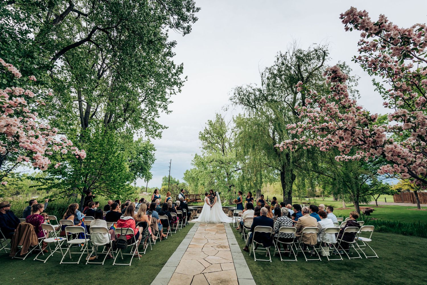 Outdoor wedding ceremony setup with rows of chairs on green lawn surrounded by blooming trees.