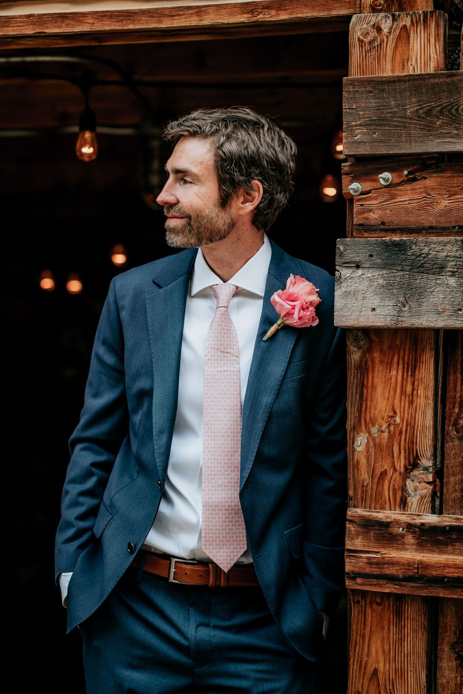 Groom in navy suit with pink tie and boutonniere stands by rustic wooden wall.