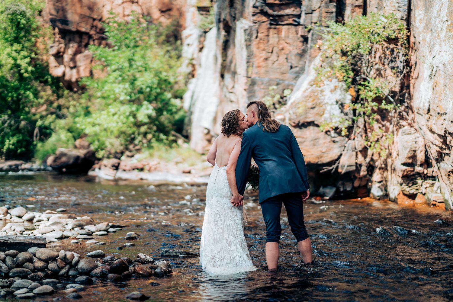 Wedding couple holding hands while walking through shallow water near scenic cliffs.
