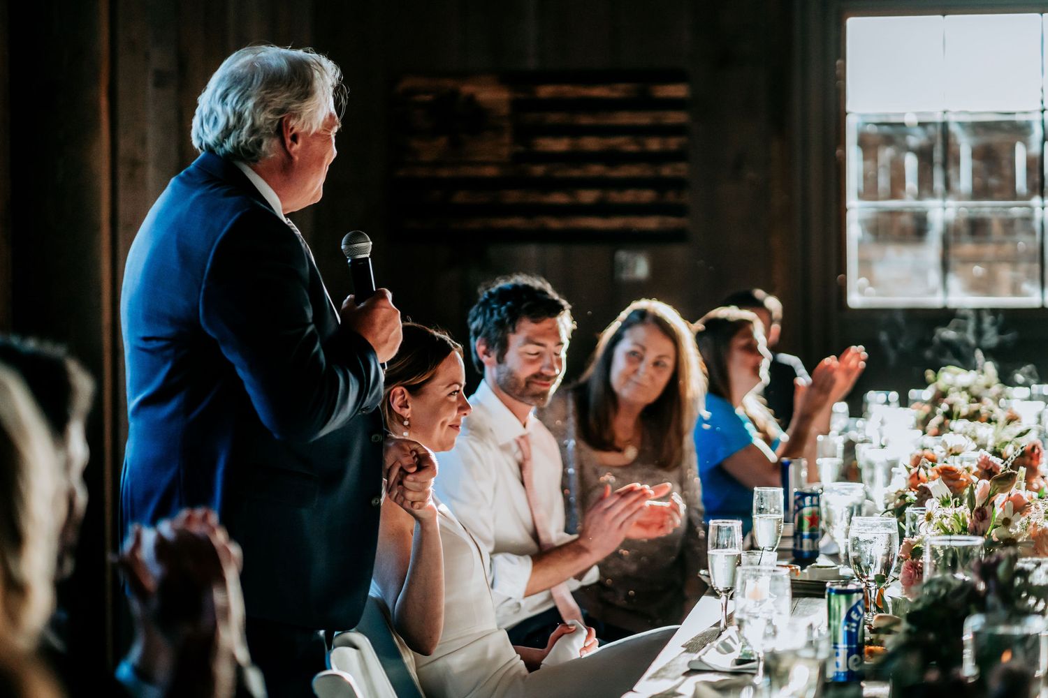 During a wedding reception toast, guests applaud and celebrate at a rustic barn venue with elegant table settings.