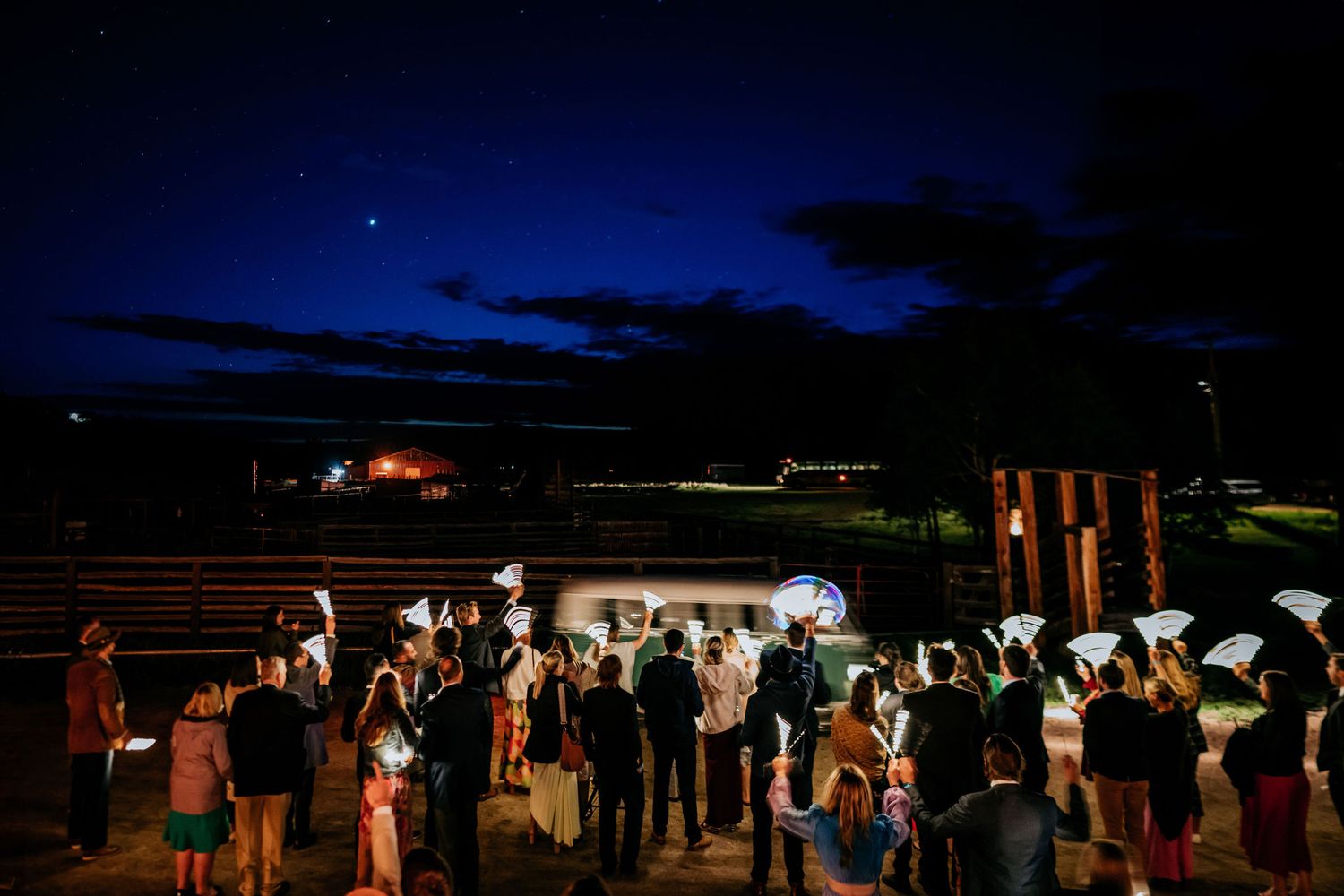 Wedding guests gather for an outdoor evening reception under a dark blue sky with twinkling lights.