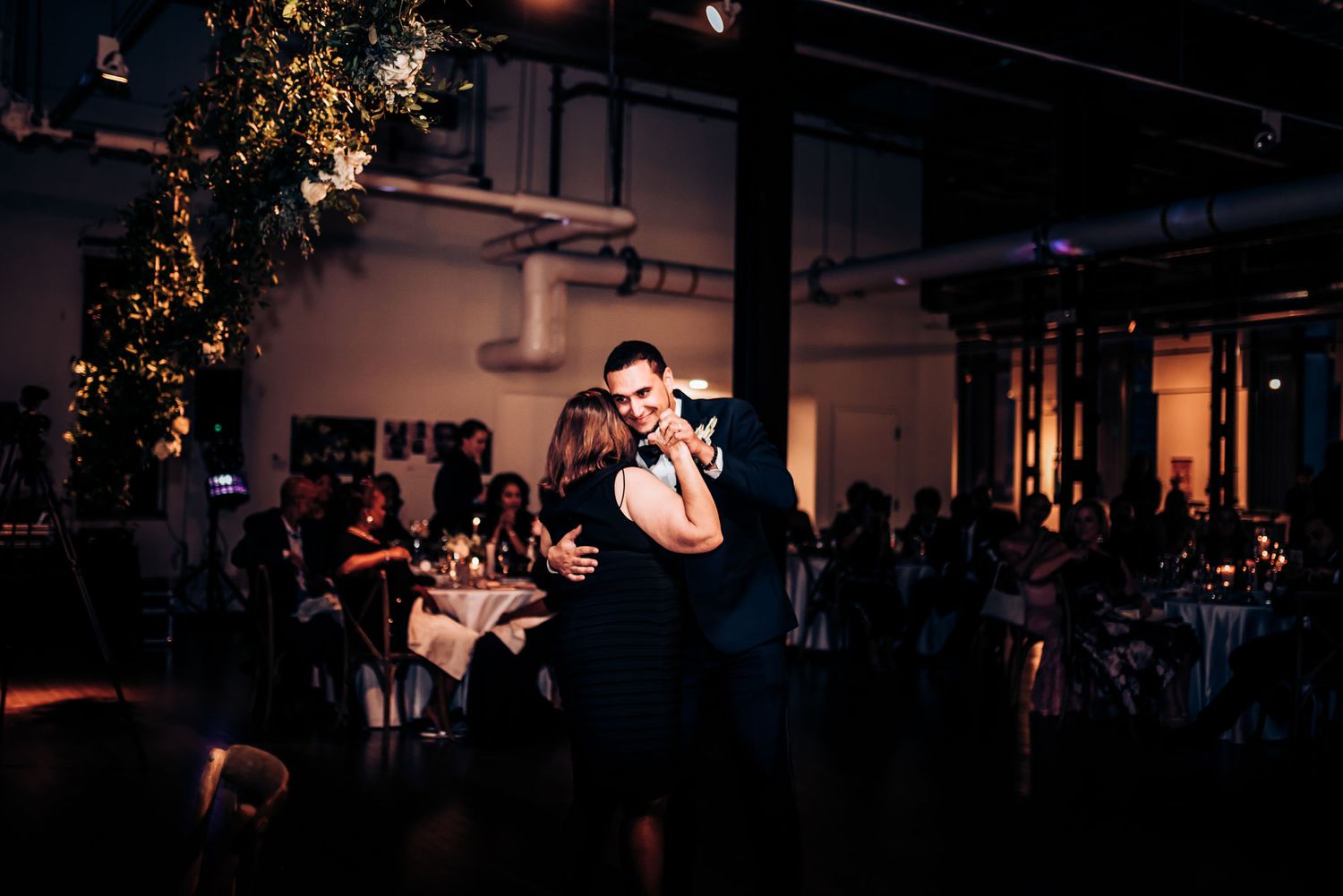 A couple dances intimately in a dimly lit reception venue with guests seated at tables in the background.