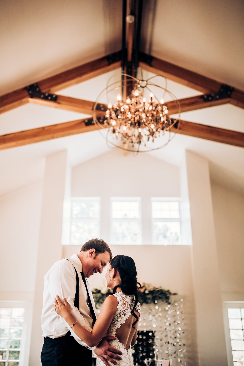 A romantic moment beneath an elegant chandelier in a bright wedding venue with wooden beams.