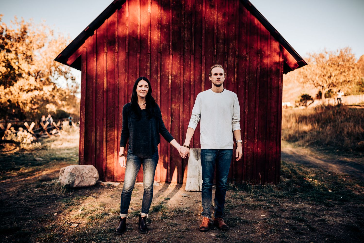A couple holding hands while standing in front of a rustic red barn at sunset.