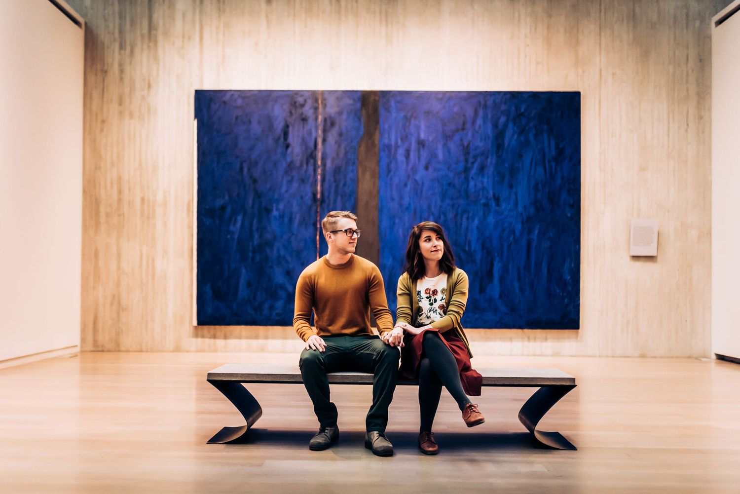 Two people sit on a modern bench in front of a vibrant blue painting in an art museum.