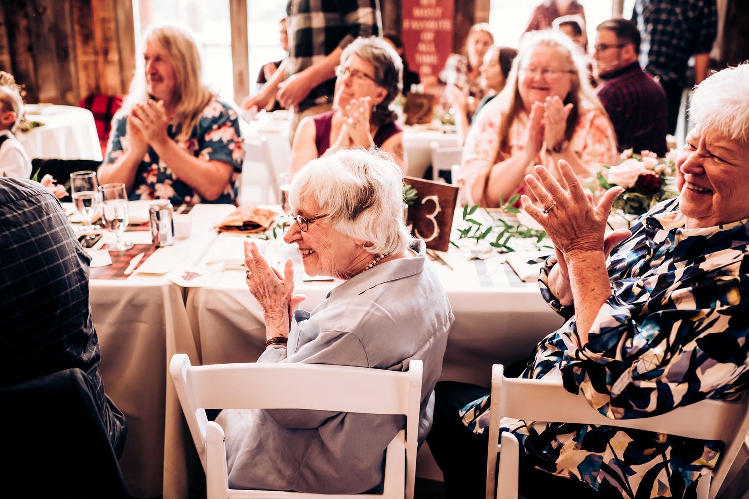 Wedding reception guests seated at round tables with white linens applauding.