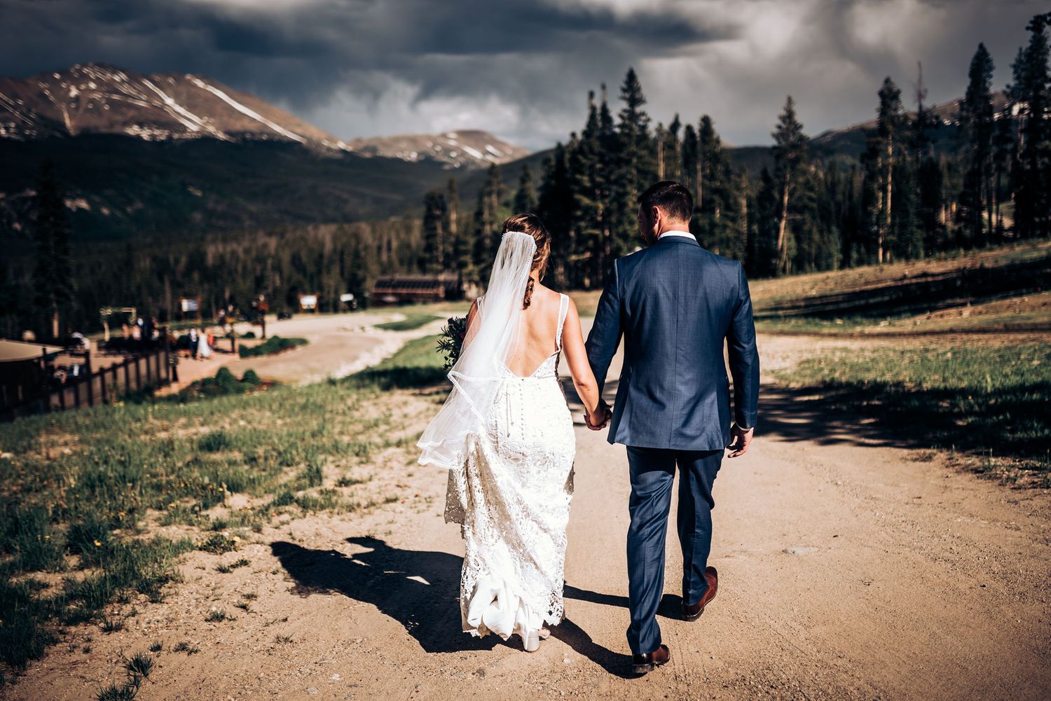 Newlyweds walk hand in hand on mountain path with scenic mountain vista.
