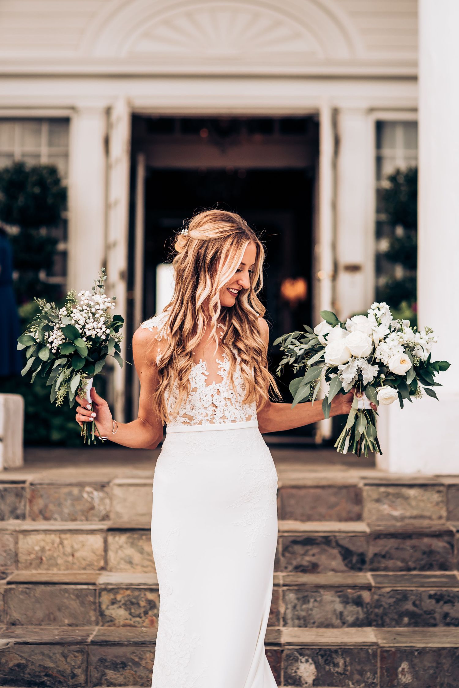 Elegant bride in lace gown holds two floral bouquets while standing on stone steps.