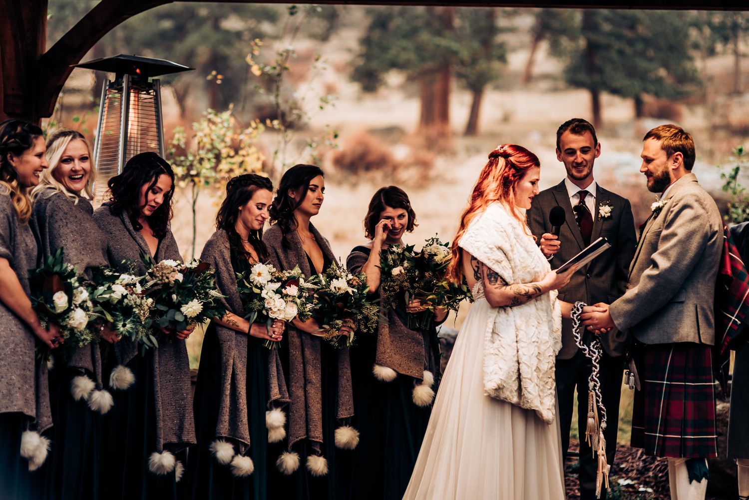 Outdoor wedding ceremony with bridal party in gray dresses and Scottish kilts against natural backdrop.