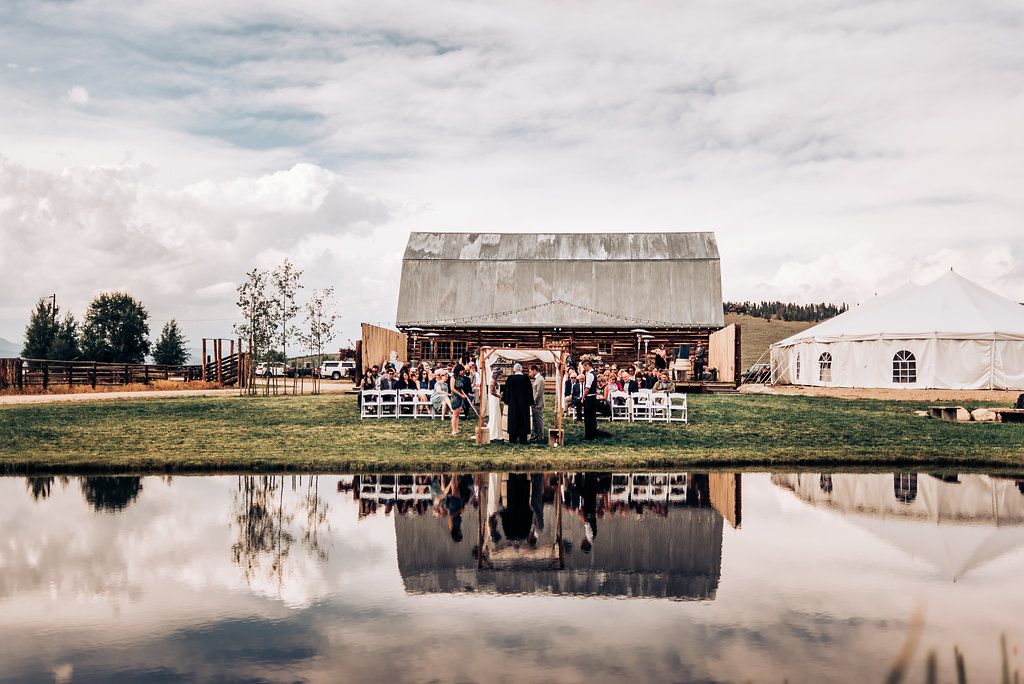 A rustic barn wedding venue reflected perfectly in a still pond on a cloudy day.