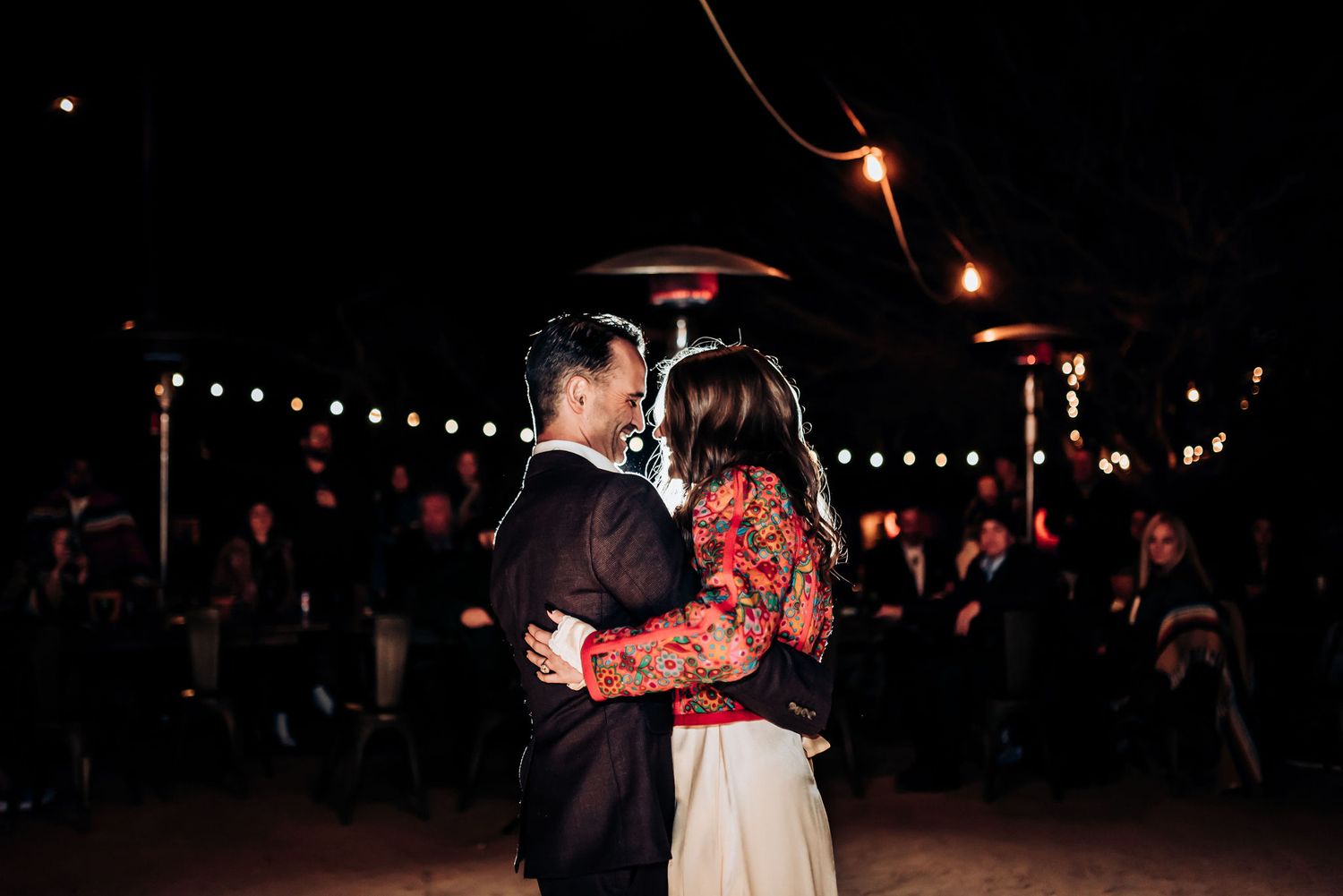 Dancing couple embraces on outdoor patio under warm string lights and heaters.