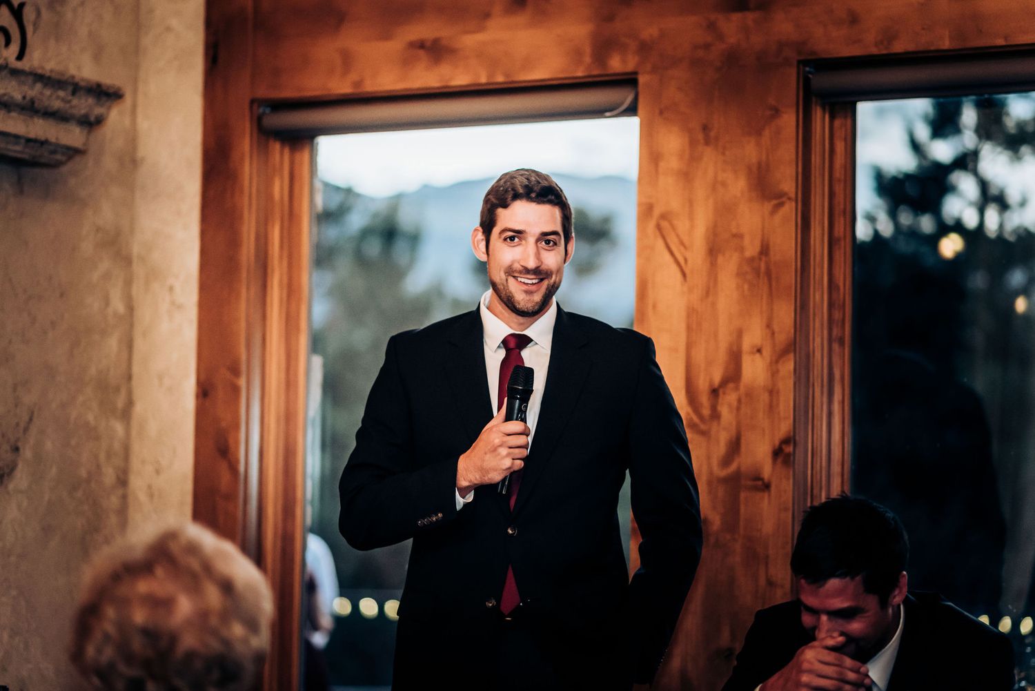 A speaker in a black suit gives a wedding toast in a rustic wood-framed venue.