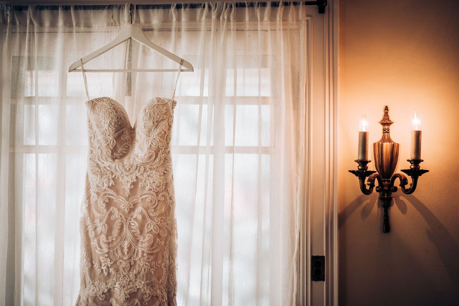 Lace wedding dress hangs by window with elegant wall sconce creating romantic ambiance.