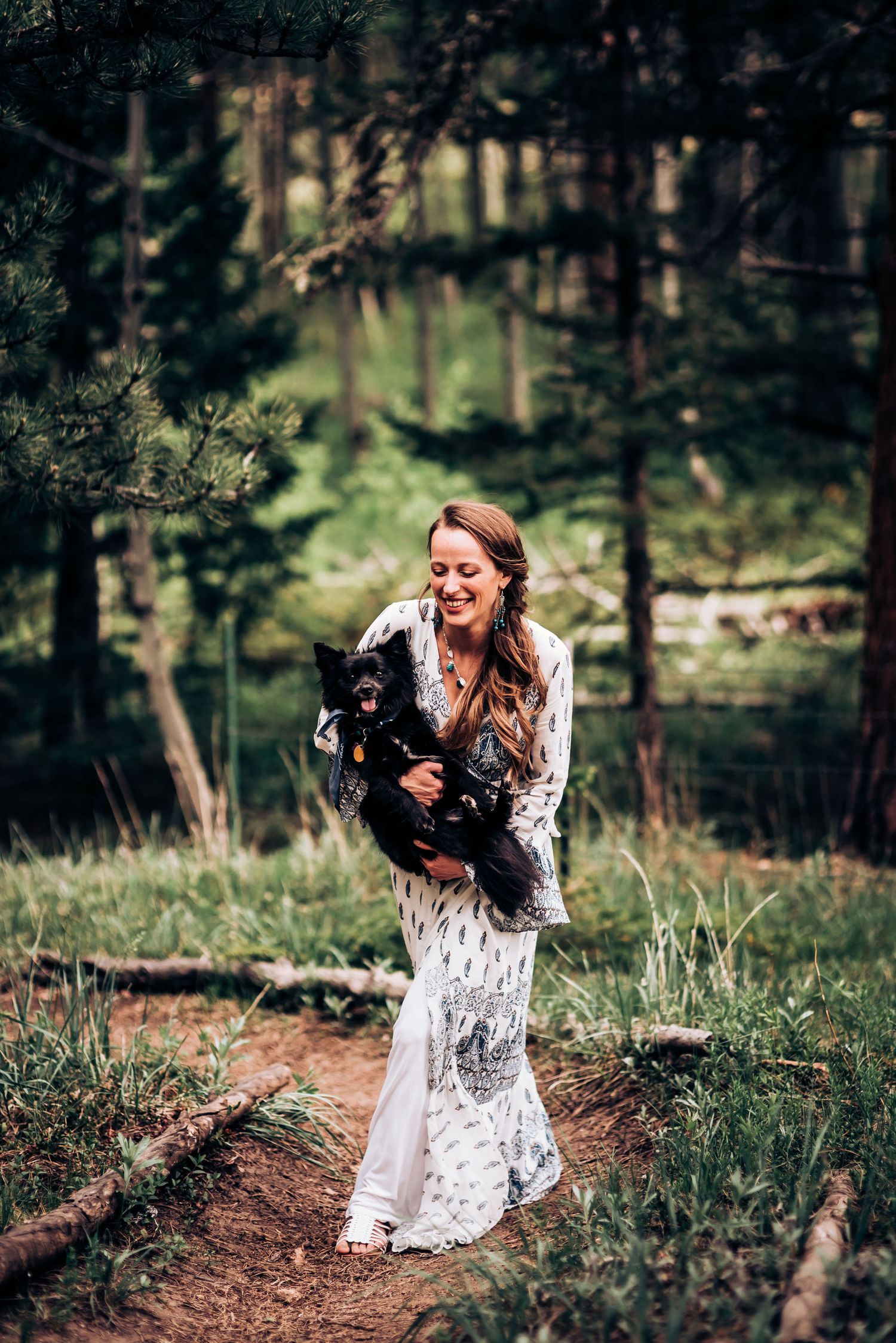 A woman in a white lace dress holds a small black dog while standing on a forest trail.