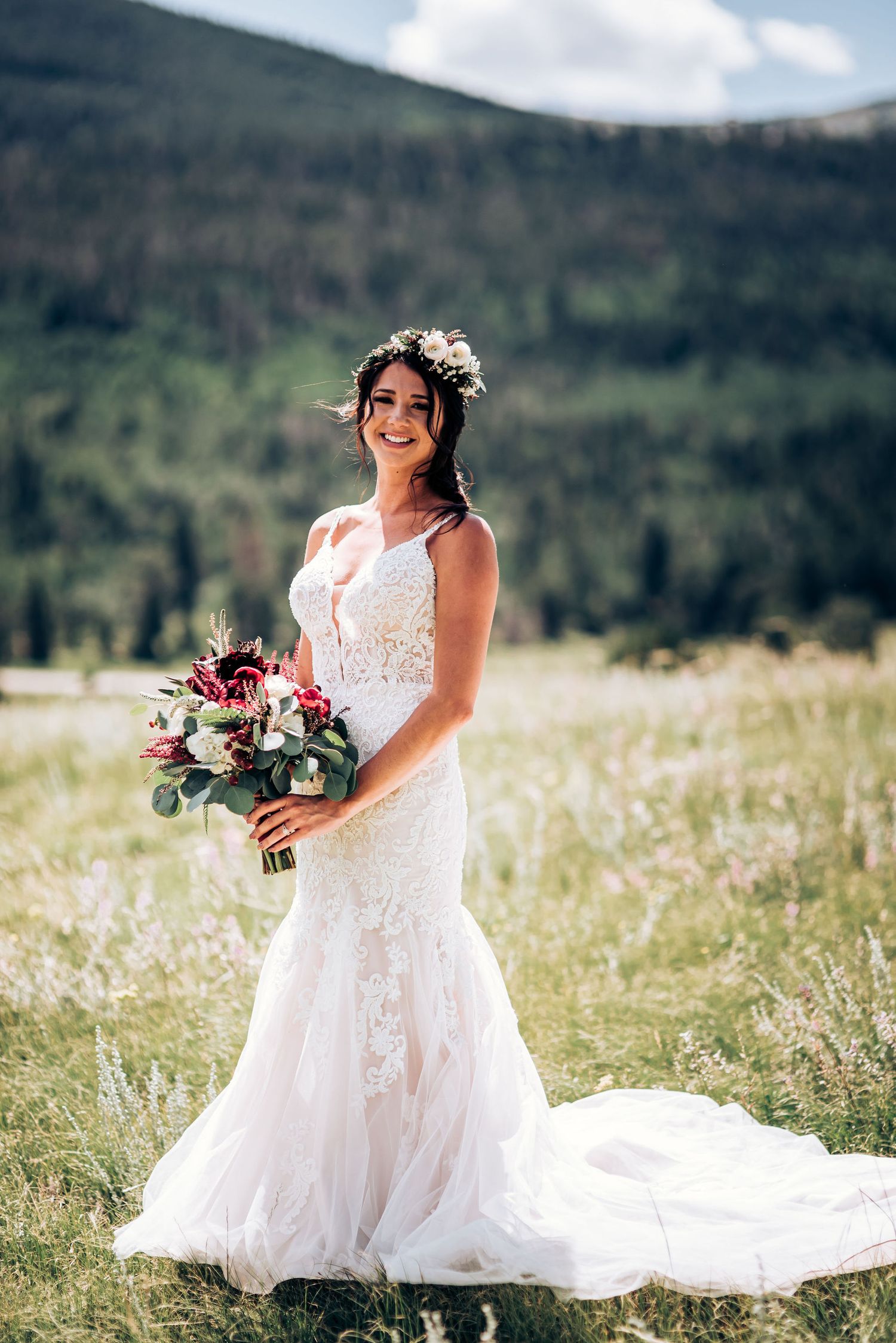 A lace wedding dress with floral details cascades across a mountain meadow landscape.