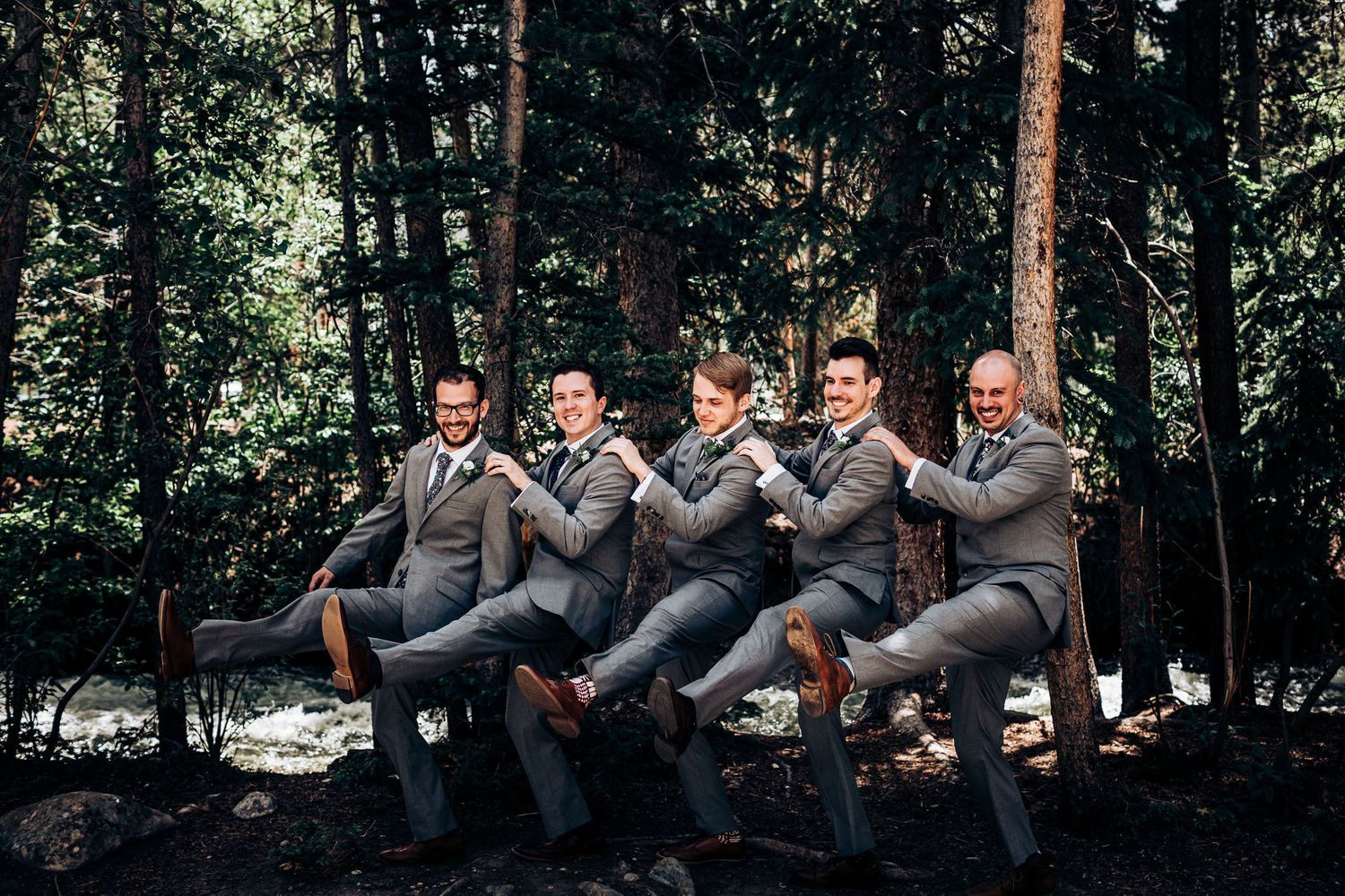 Groomsmen in gray suits strike playful synchronized kicking pose in wooded setting.