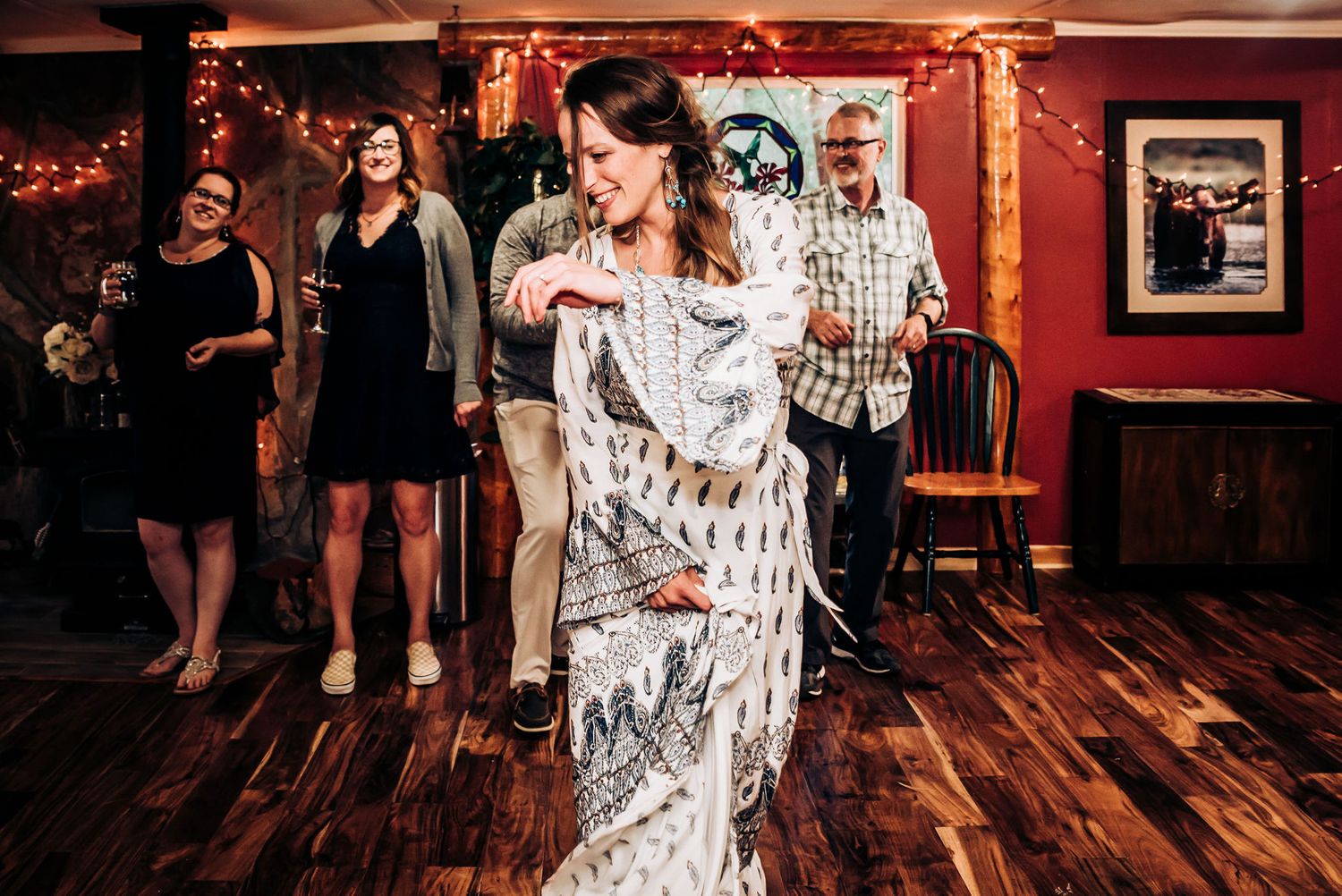 Partygoers dance on hardwood floor in rustic venue decorated with string lights.
