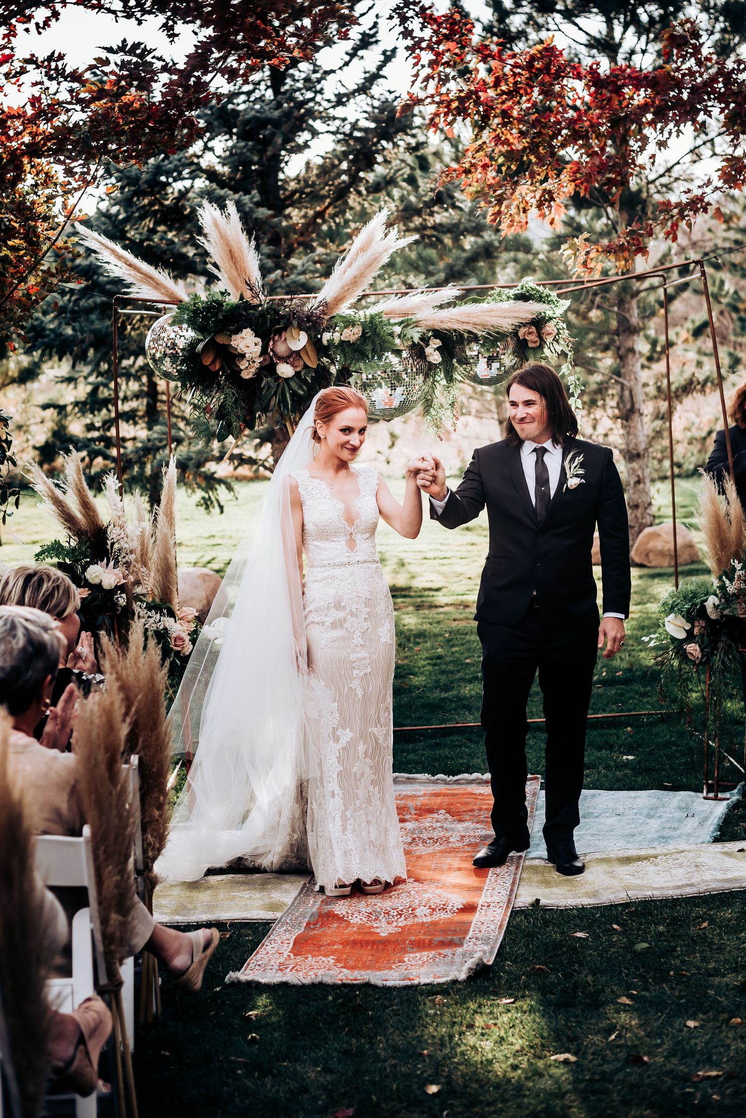 Ceremony under a boho floral arch with pampas grass on an outdoor path surrounded by autumn foliage.