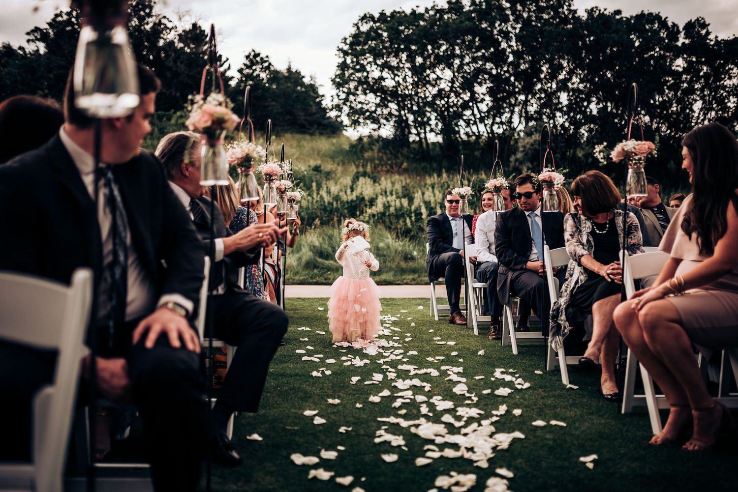 Wedding guests hold champagne glasses during outdoor ceremony with flower petals scattered on grass.