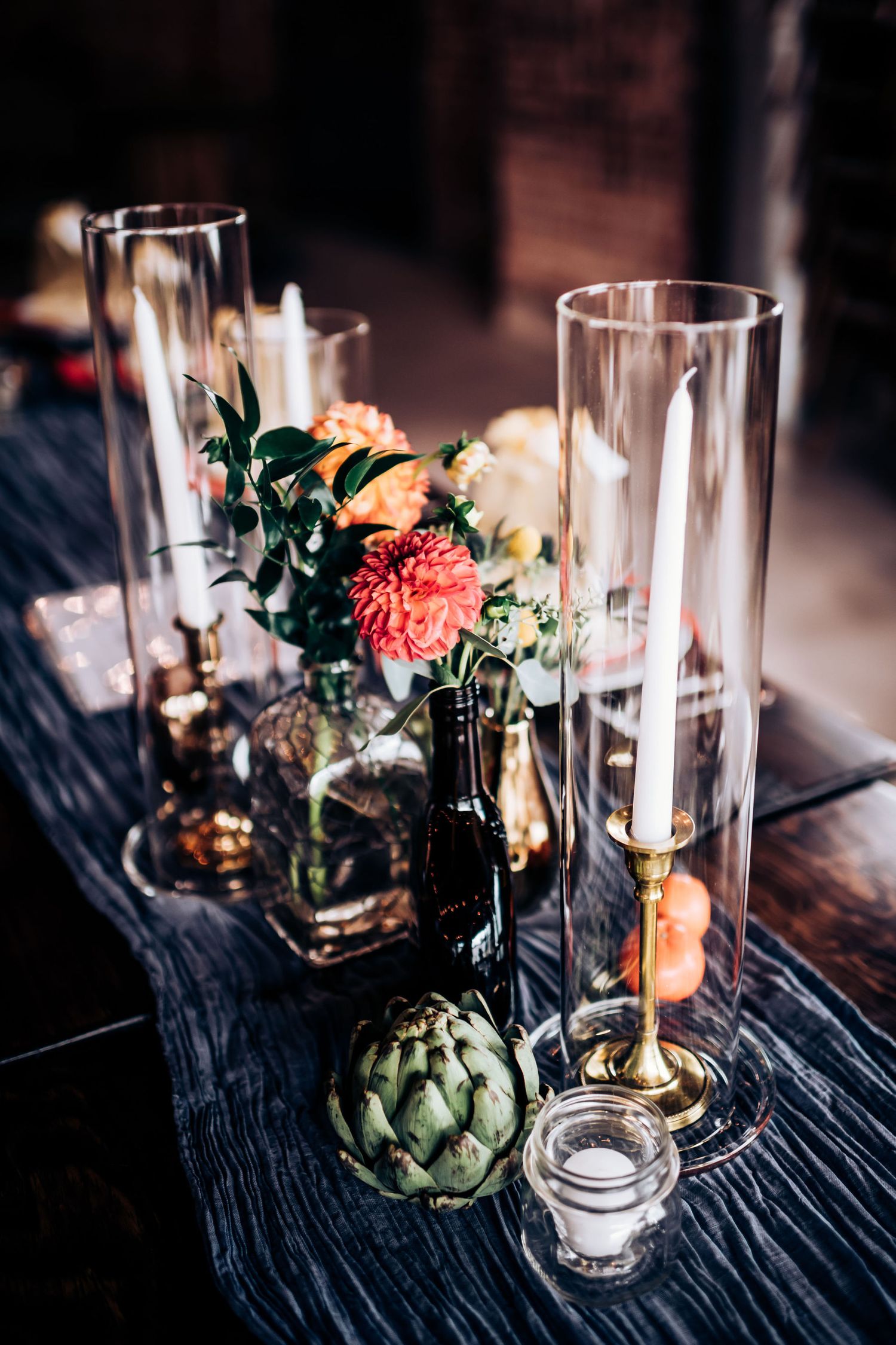 Artichoke and floral centerpiece with tall glass candleholders on dark wooden table.