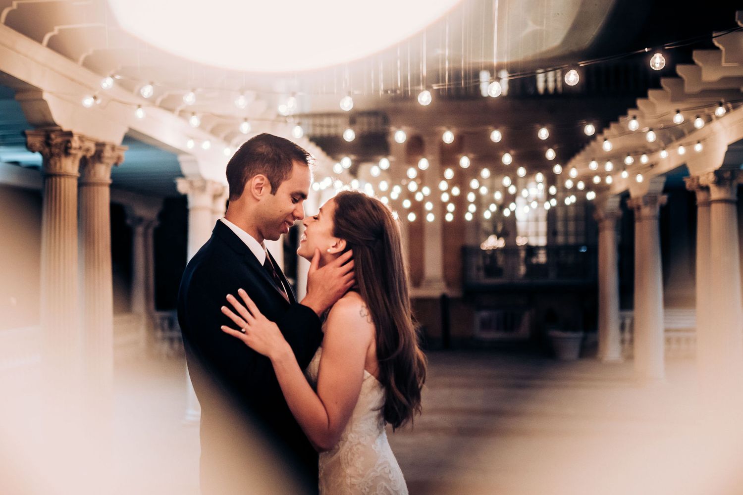 A couple embraces under romantic string lights and classical columns during their wedding celebration.