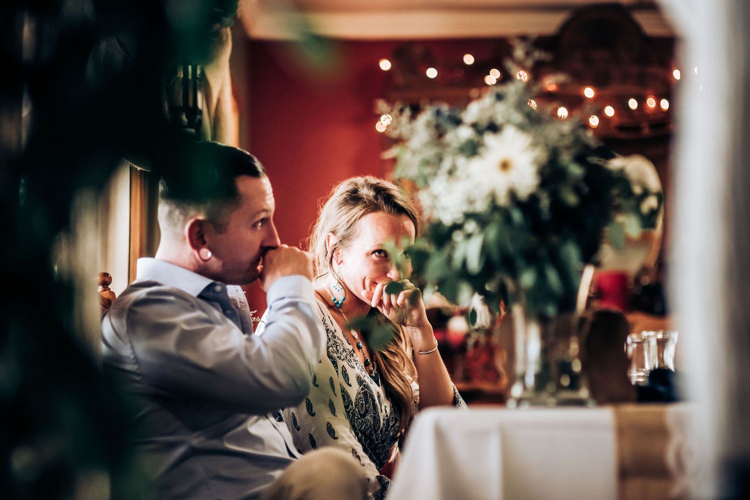 Emotional wedding reception scene captured through foliage with string lights creating warm ambiance.