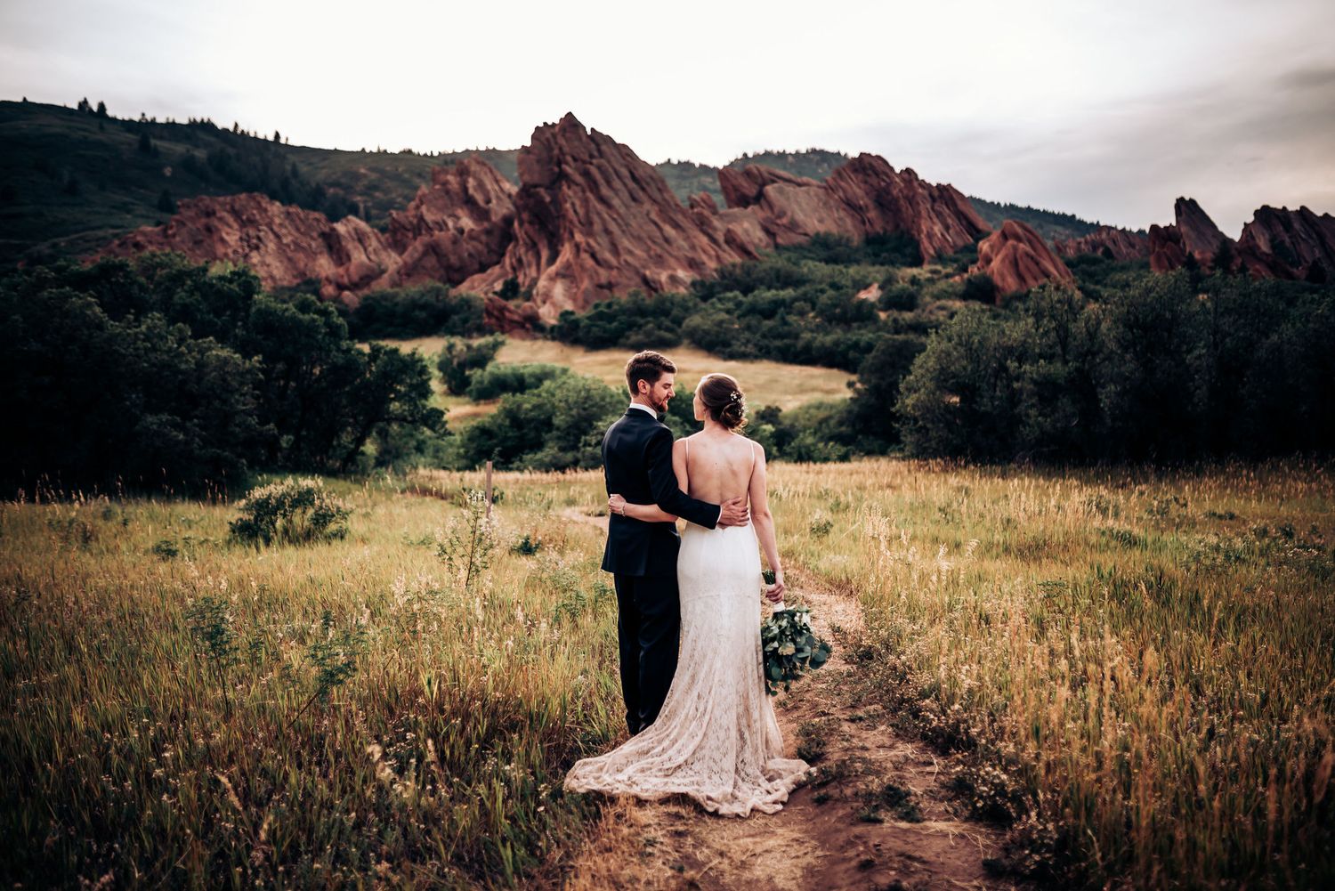 A wedding couple stands on a mountain trail overlooking dramatic red rock formations.
