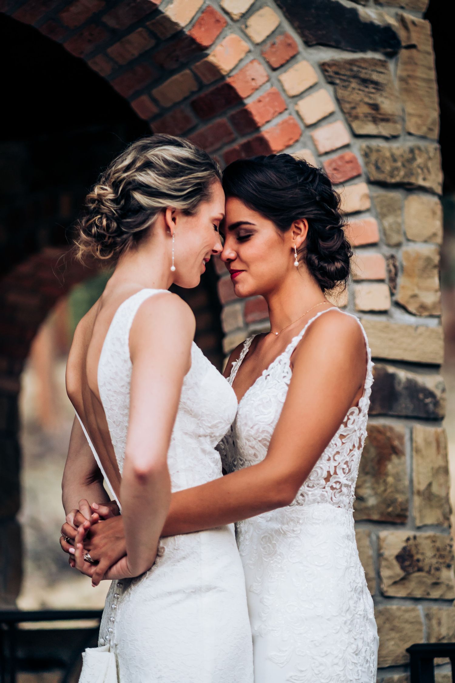 Two people in white lace wedding dresses share an intimate moment against a brick archway.
