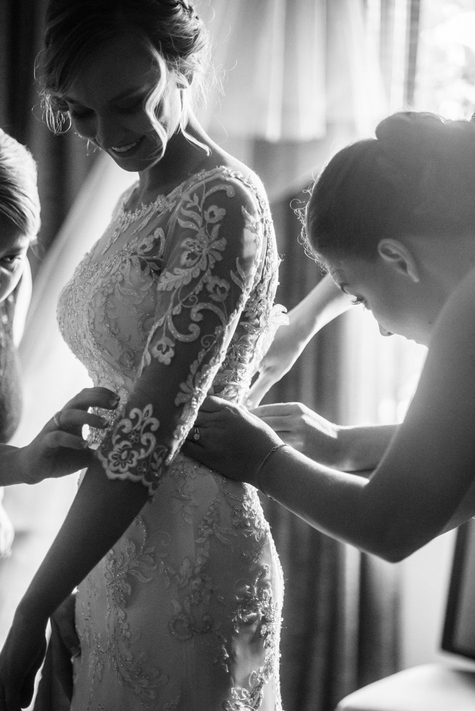 Black and white photo capturing final adjustments to a lace wedding gown during bridal preparations.