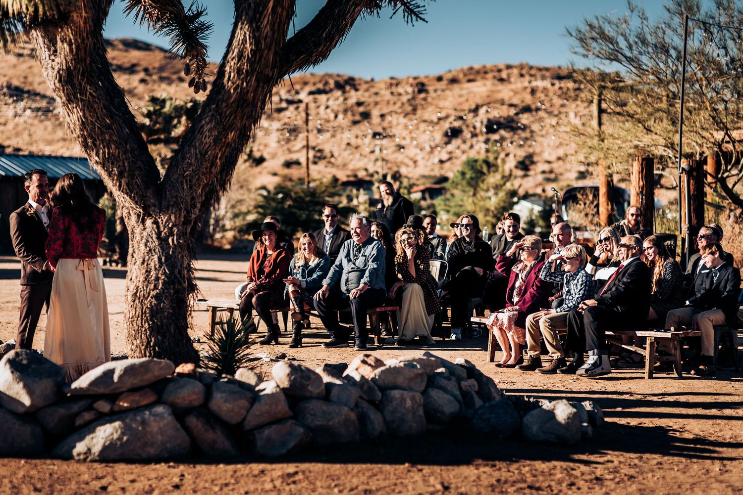 Outdoor desert wedding ceremony with guests seated under a large tree against rocky terrain.