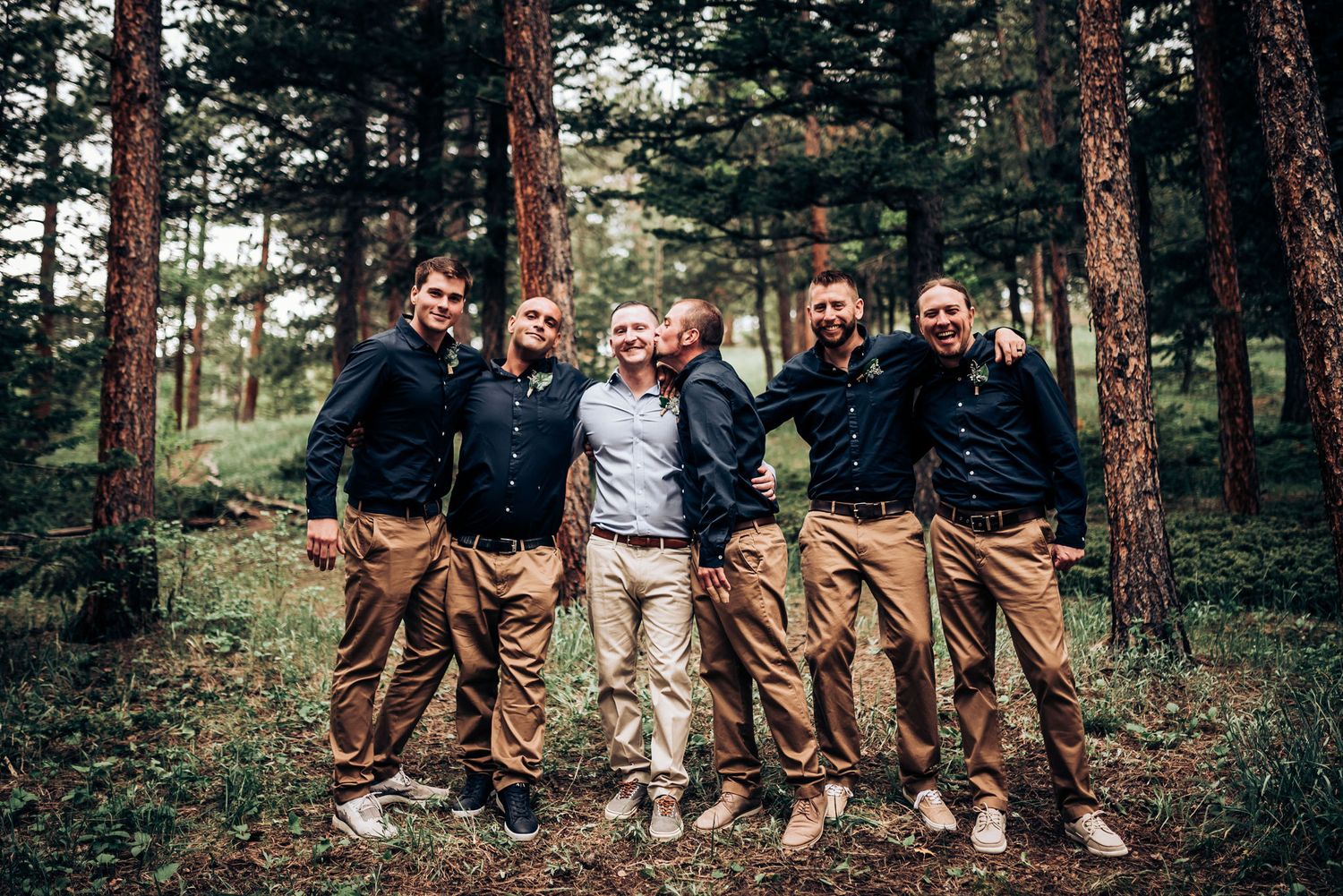Group of groomsmen in matching navy shirts and khakis pose together in forest setting.