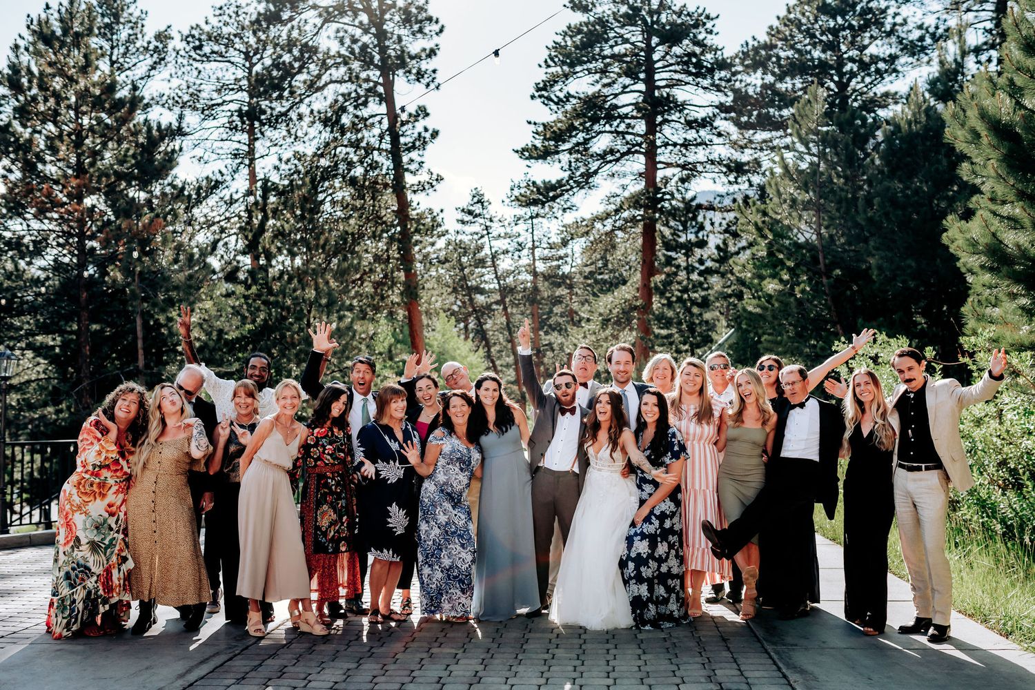 Wedding group portrait with guests in mixed formal attire against forest backdrop.