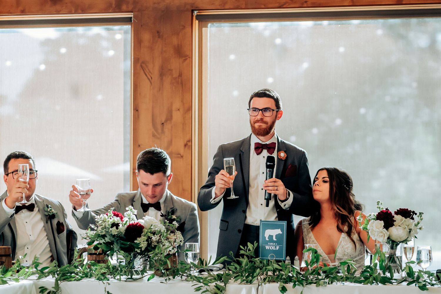 Wedding reception toast during speeches at the head table adorned with floral arrangements.