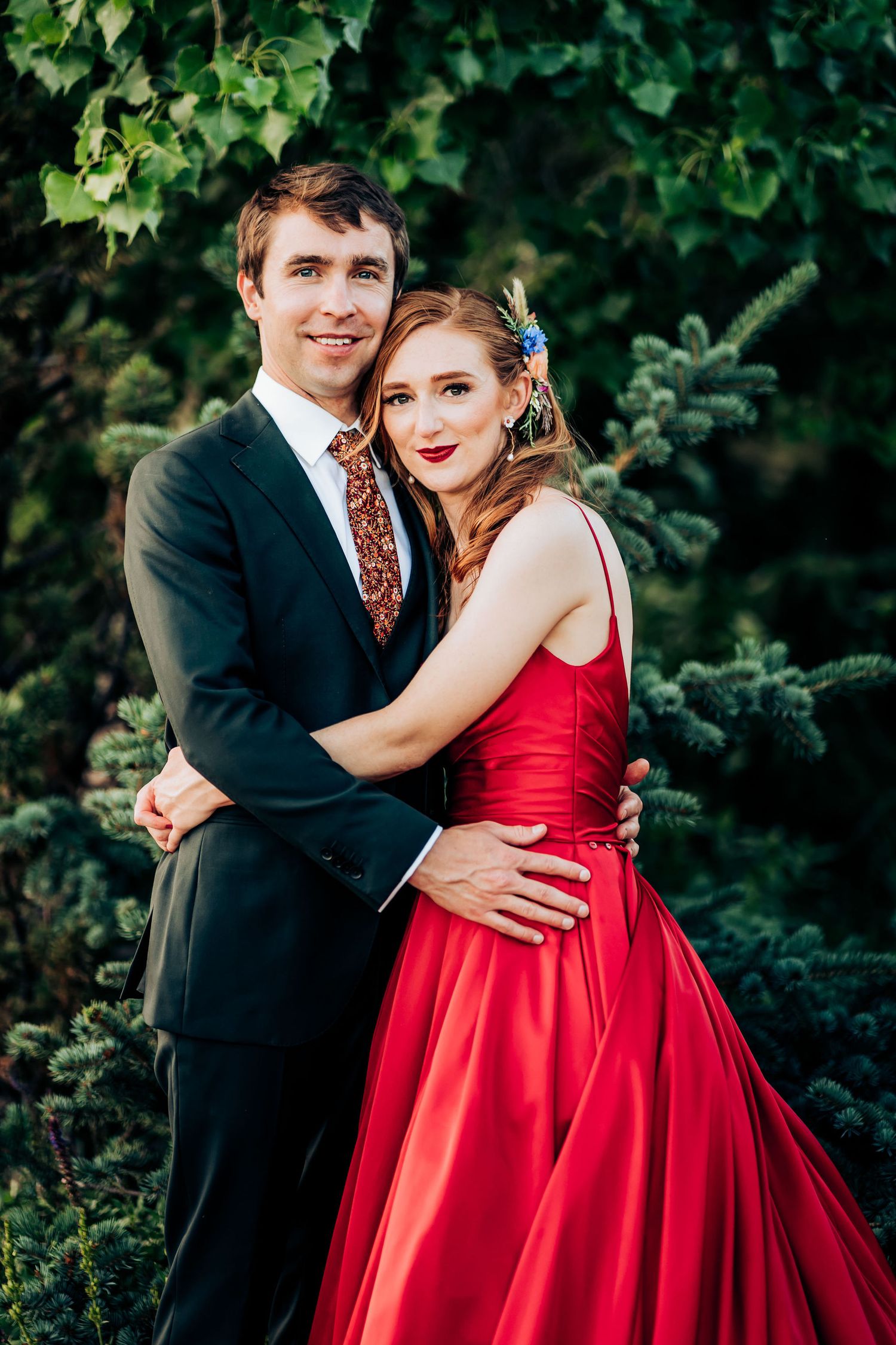 A couple poses against greenery, with her in a red ballgown and him in a black suit with patterned tie.