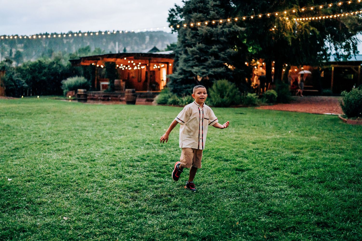 Child running playfully on grass with string lights and buildings in background at dusk.