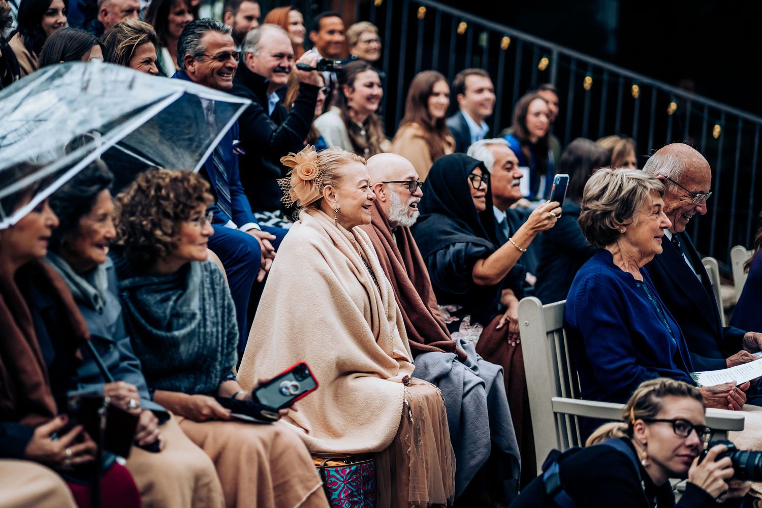 Wedding guests watching ceremony while holding clear umbrellas in outdoor seating area.
