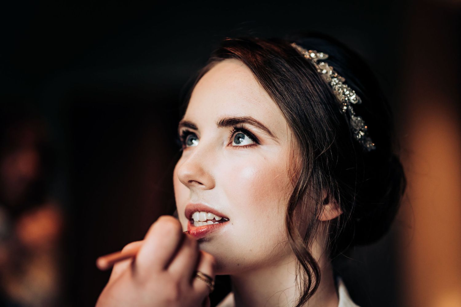 Professional makeup artist applies lipstick during wedding preparation in a dramatic low-light setting.