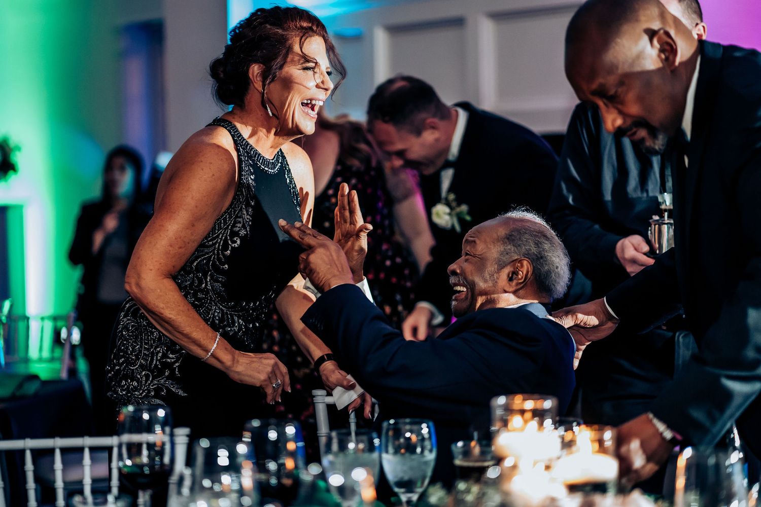 Wedding guests interact enthusiastically during a reception with colored mood lighting.