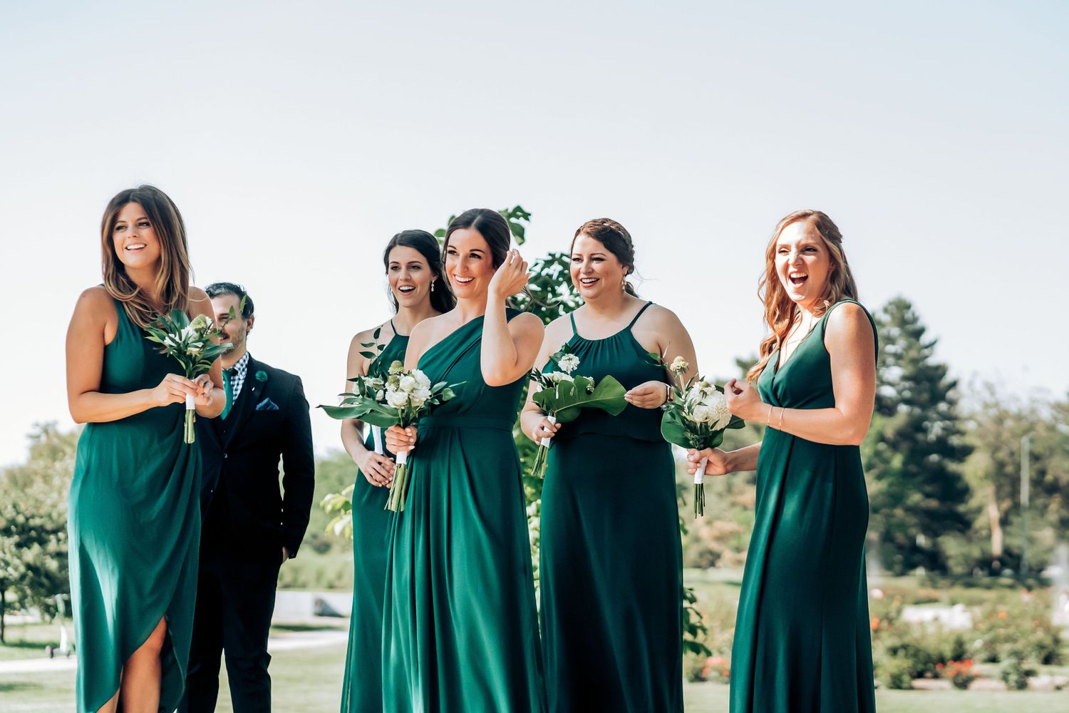 Bridesmaids in elegant emerald green dresses stand together holding bouquets outdoors.