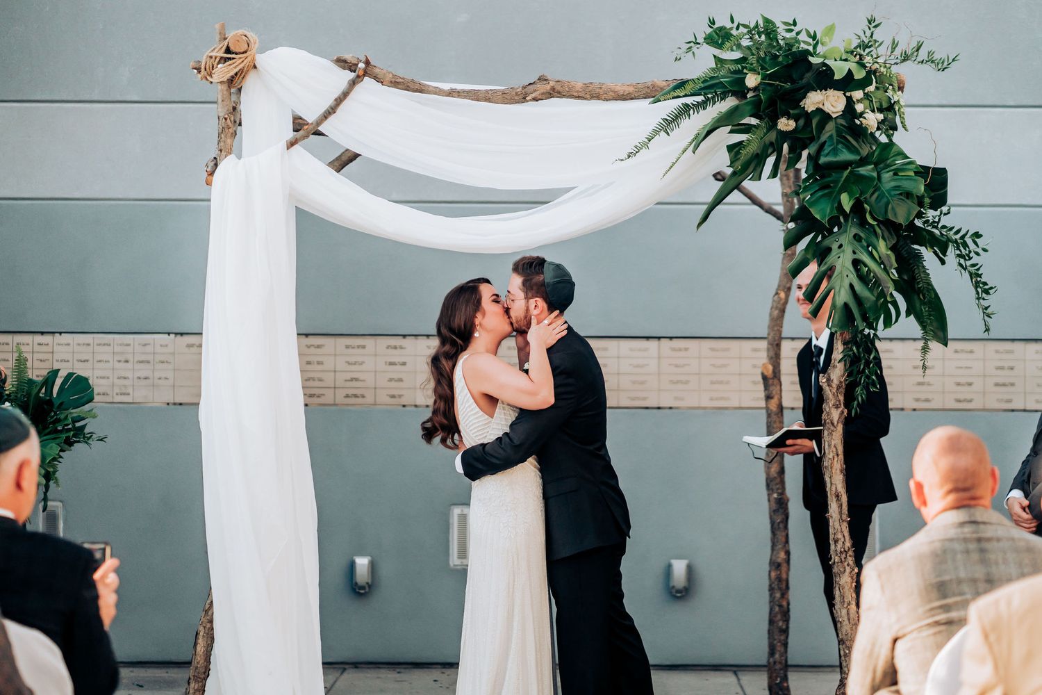 Couple sharing first kiss under white draped wedding arch with greenery accents.