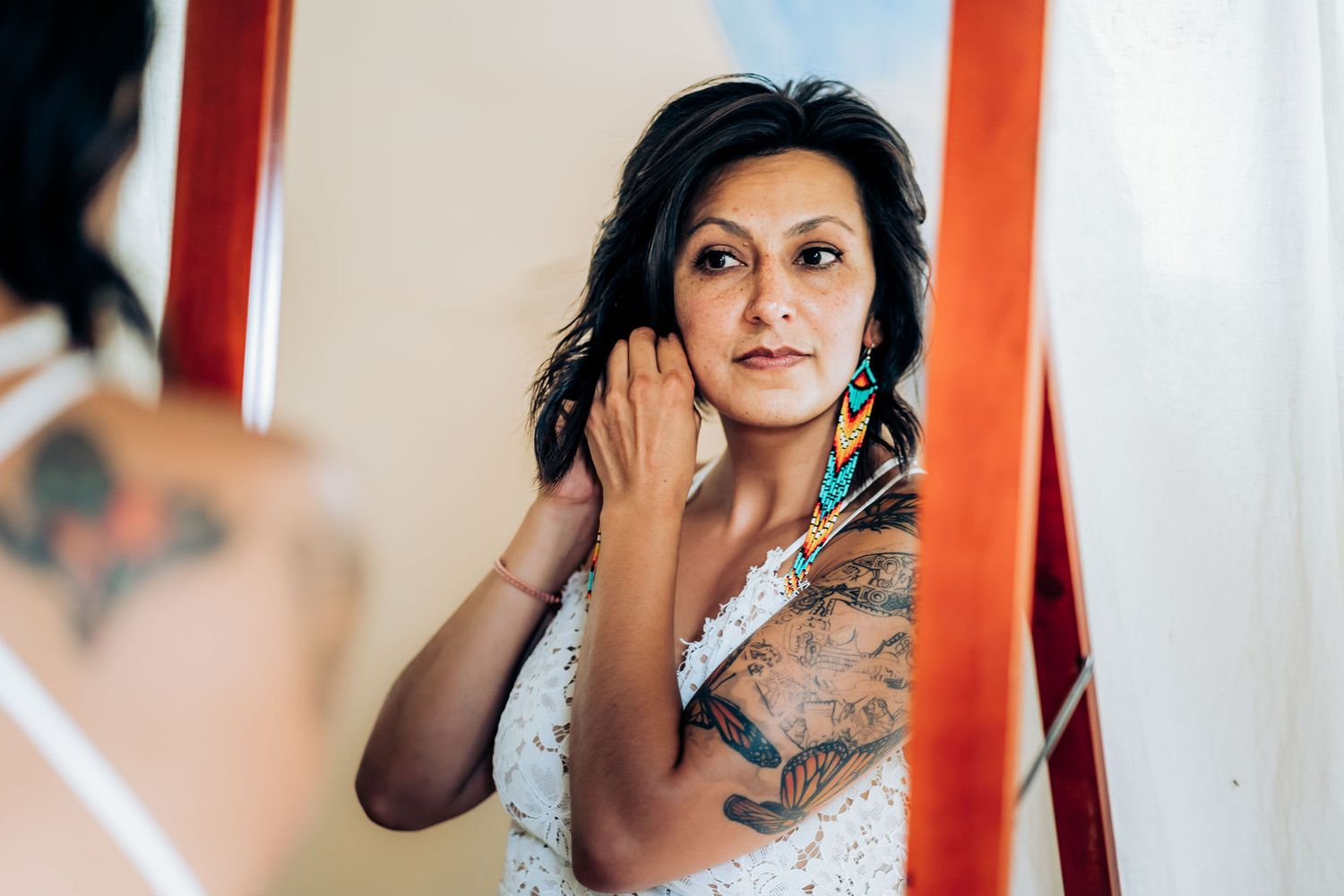 Person in white lace dress with tattoos putting on earrings while looking in mirror.