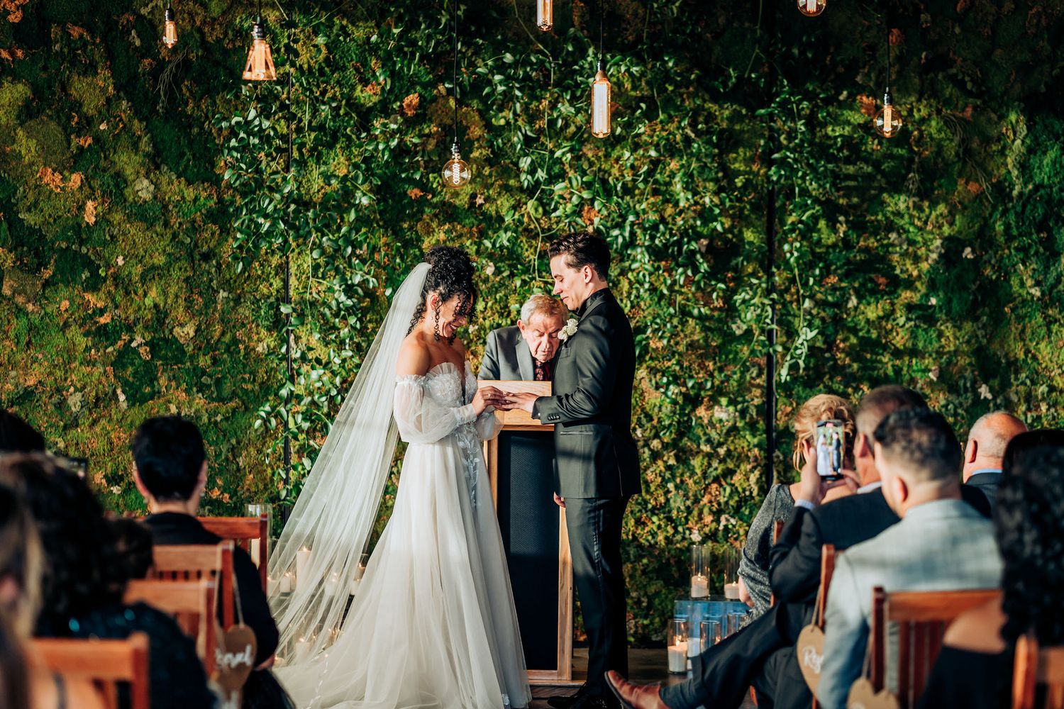 Wedding ceremony under string lights and ivy-covered walls with guests seated.