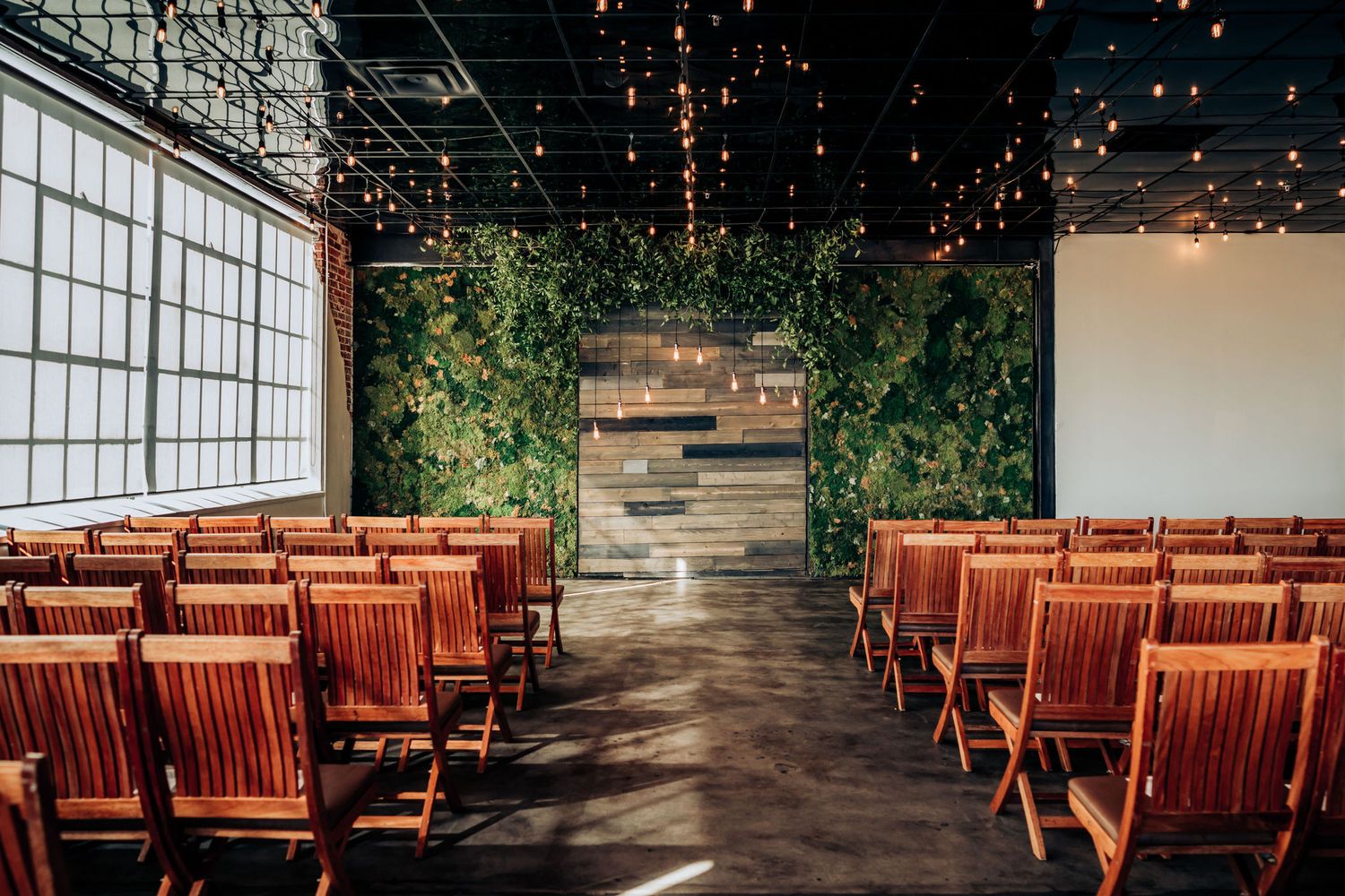 Modern wedding ceremony space with wooden chairs, string lights, and lush greenery wall decorations.