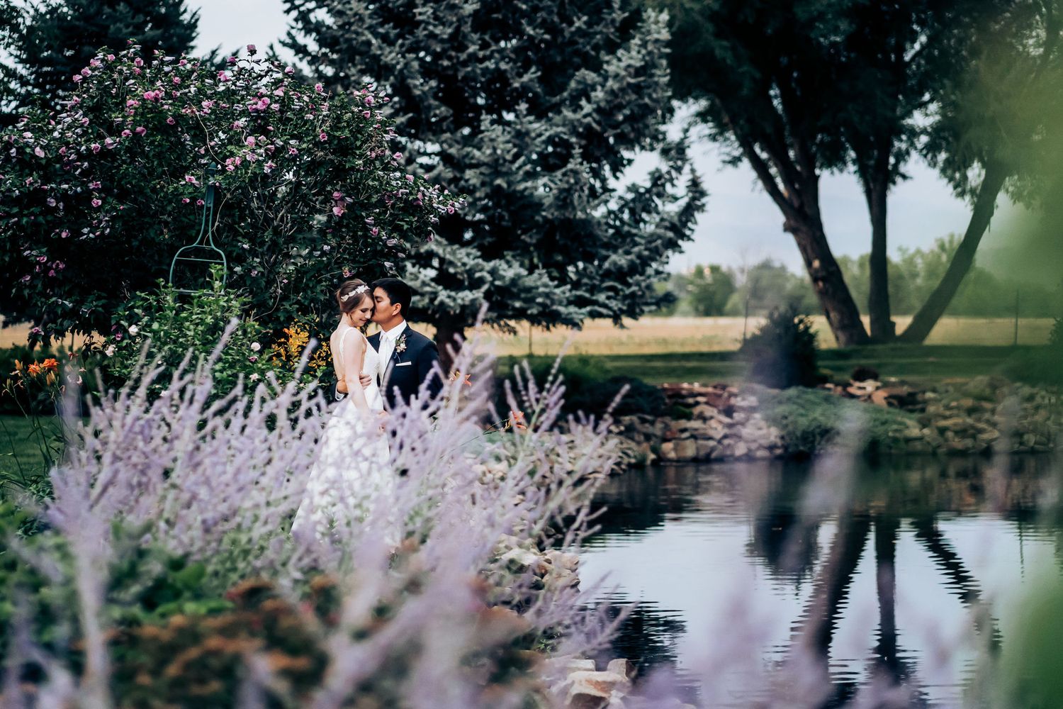 Romantic wedding couple embraces near a serene pond with purple wildflowers in the foreground at sunset.