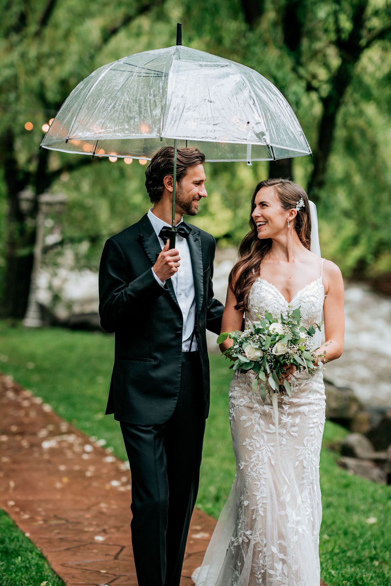 Newlyweds laugh together while holding a clear umbrella during their outdoor wedding photoshoot.