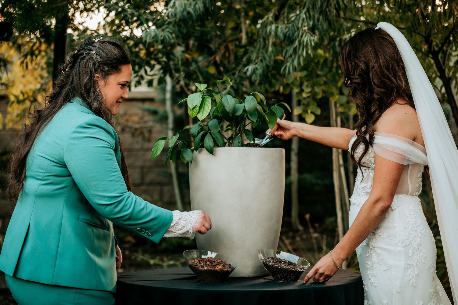 Two brides laughing together while performing a tree planting unity ceremony at their outdoor wedding.