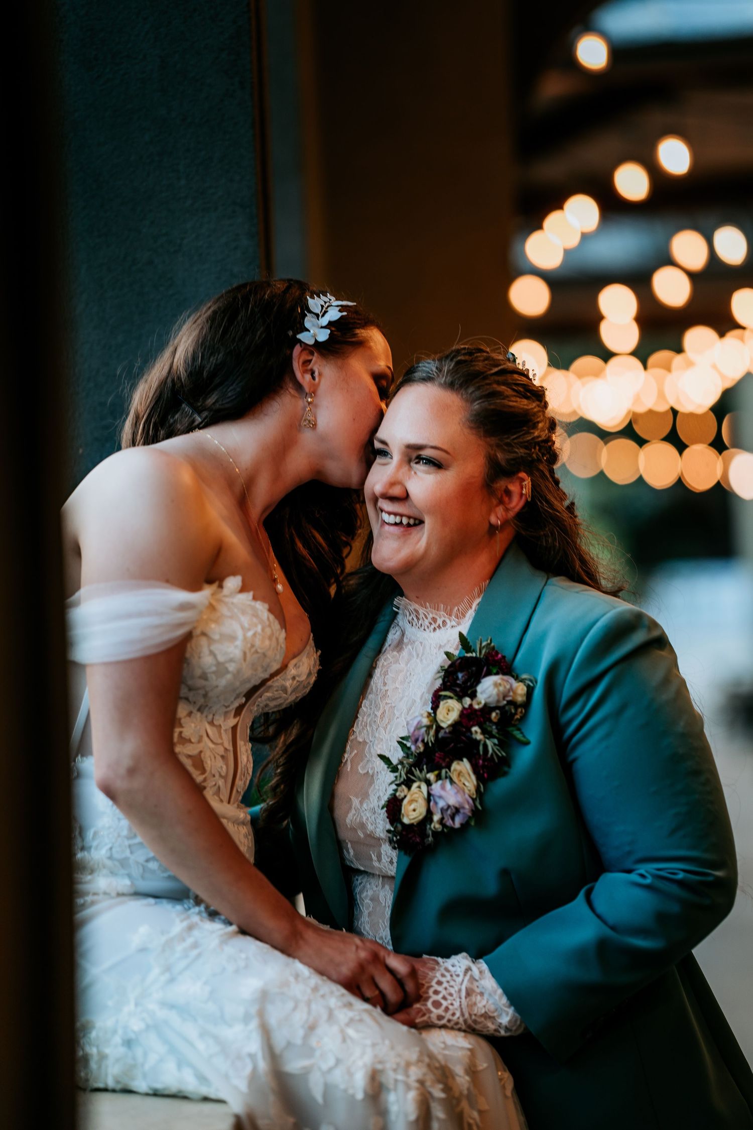 Two brides share an intimate moment indoors, one whispering to the other amid warm bokeh lights.