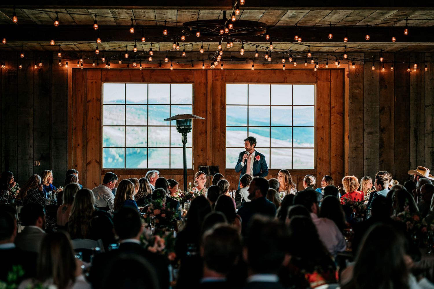 Wedding speaker addresses a crowd in rustic venue with large windows overlooking ocean view.