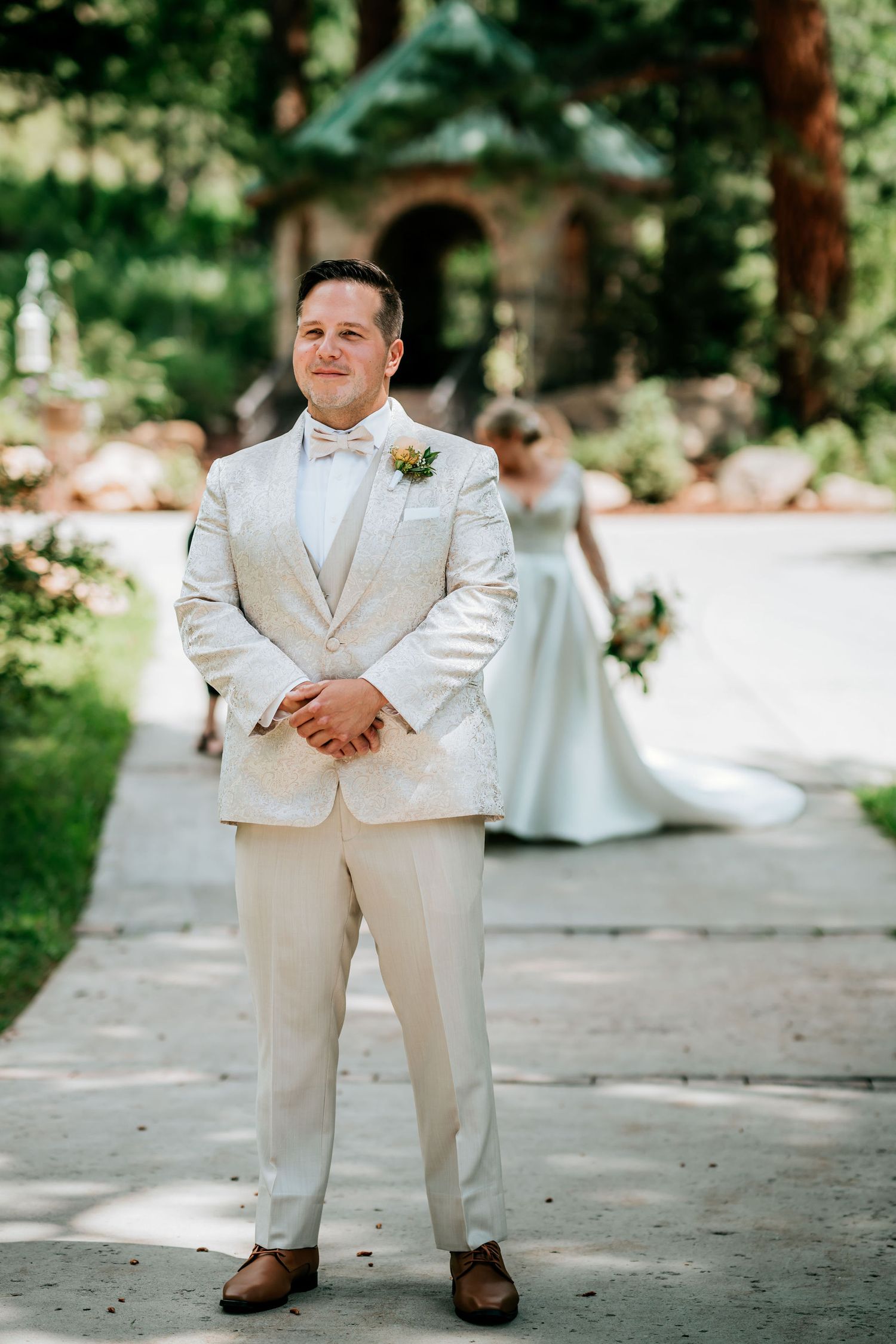 A groom in a cream-colored suit stands waiting on a stone path with garden scenery behind him.