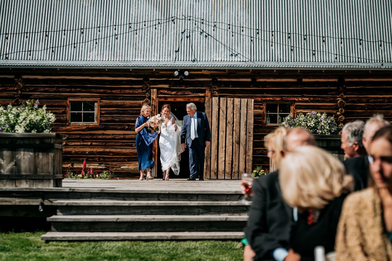Wedding guests gather outside rustic wooden lodge with steps and planters.